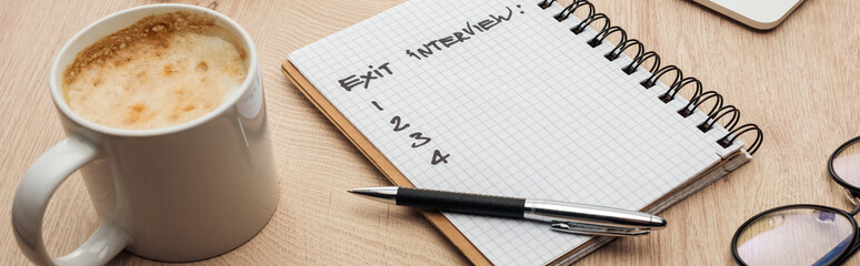 panoramic shot of notebook with exit interview lettering and numbers on wooden table with pen, glasses and cup of coffee