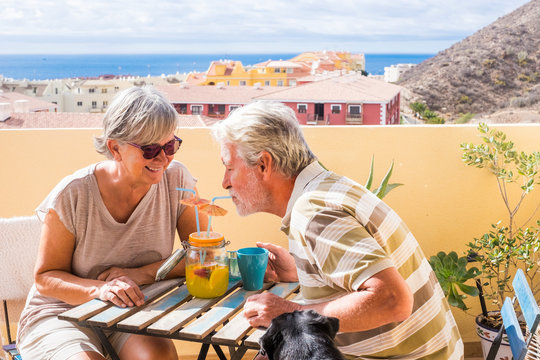 elderly senior couple man and woman with nice dog pug on the terrace. drink together from the same vase a fruit juice for healthy lifestyle. summer nice time of vacation in a terrace and ocean view