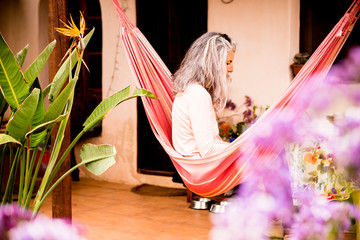 Old beautiful grey white hair woman sit down outdoor on hammock enjoying the coloured home - defocused violet flowers on foreground and relaxing concept