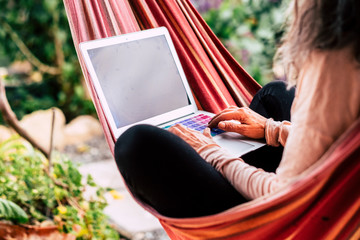Old lady working on a computer laptop sitting on a coloured hammock - job and work everywhere with...