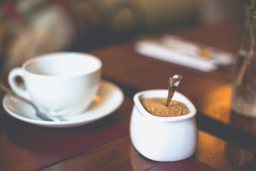 Close-up white sugar bowl with brown sugar on a white cup with a saucer on the table in a restaurant