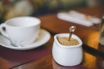 Close-up white sugar bowl with brown sugar on a white cup with a saucer on the table in a restaurant