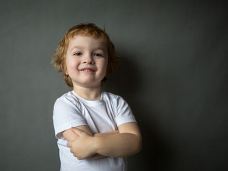 Cute cheerful happy curly-haired boy stands, hands clasped  and looking in to the camera. Close-up. Gray background.