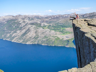 A girl wearing orange hoodie sitting at the edge of a steep cliff of Preikestolen, with legs hanging down. A view on Lysefjorden. Fjord goes far inland. Girl enjoys the view, feeling free and happy.