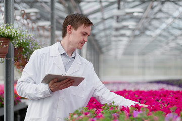 A young man in a white coat, a scientist biologist or agronomist examines and analyzes the flowers and green plants in the greenhouse.