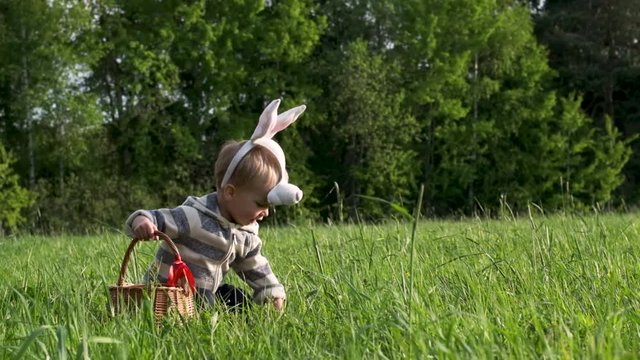 Cute little kid in bunny mask searching for easter eggs on lawn in nature or park. Easter egg hunt. Slow motion.