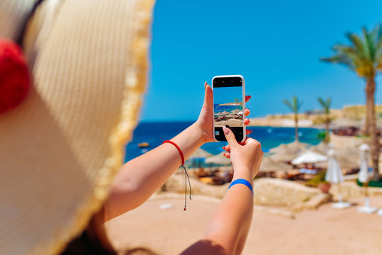 Young travelling woman blogger in hat taking photos of summer sunny sea with smartphone camera. Brunette girl making photography on summer vacation