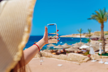 Young travelling woman blogger in hat taking photos of summer sunny sea with smartphone camera. Brunette girl making photography on summer vacation