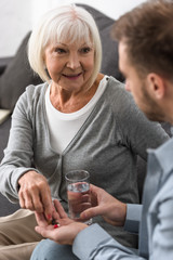 cropped view of man giving to senior mother glass of water and pills