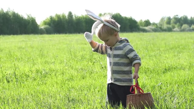 Cute little child on Easter day. Toddler boy with bunny ears hunts for Easter eggs on the lawn in nature or park. Slow motion.