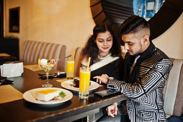 Lovely indian couple in love, wear at saree and elegant suit, sitting on restaurant. On table sweet desserts and juices. Man looking at his watches.