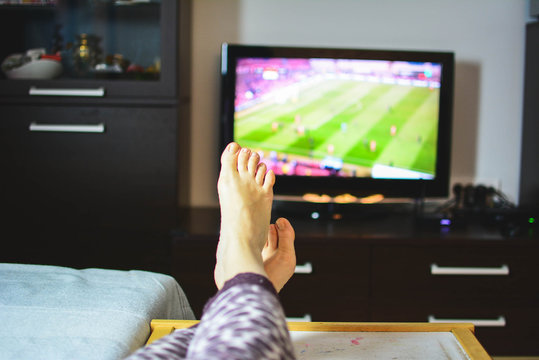 A woman with her legs stretched out on a small table in the living room watching a football game on TV. sports and entertainment concept.