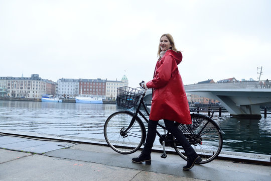Denmark, Copenhagen, happy woman pushing bicycle at city harbor in rainy weather