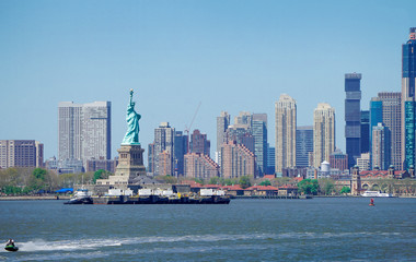 Panorama view of The Statue of Liberty in foreground and New York City skyscrapers in background, Boats in river with blue sky.