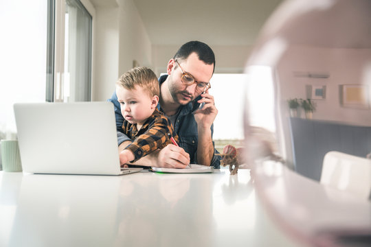 Busy father working at table in home office with son sitting on his lap