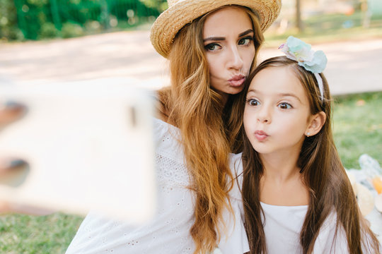 Long-haired elegant woman and funny joyful girl making selfie wih kissing face expression. Attractive young mom fooling around with daughter and taking picture of it.
