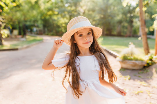 Joyful little lady with long light-brown hair posing in park in sunny warm day. Outdoor portrait of pretty dark-eyed girl in cute white dress dancing on the street enjoying summer vacation.