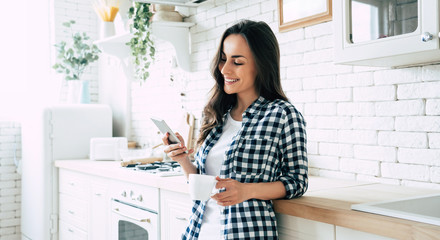 Beautiful cute smiling woman is using smart phone on the kitchen at home.
