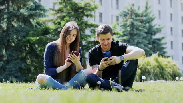 Happy friends use their phones while relaxing in a city park on the grass, chatting, surf iternete