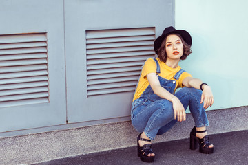 Fototapeta na wymiar young woman in blue denim overalls and yellow tshirt with black hat sensual looking at camera while posing near industrial building . outdoor shot in the summertime