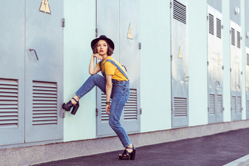 young woman in blue denim overalls and yellow tshirt with black hat sensual looking at camera while posing near industrial building . outdoor shot in the summertime