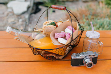 Iron basket with bottle of orange juice and sandwiches standing on wooden table. Outdoor photo of meal for picnic, empty glass and camera.