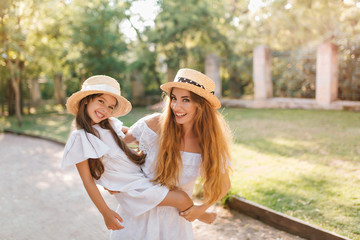 Inspired woman in good mood holding daughter and laughing while posing in alley with green trees. Outdoor portrait of adorable young mom carrying girl in white dress with cute smile.