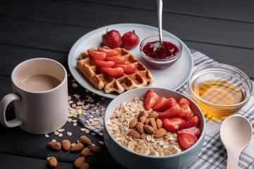 Fresh breakfast. Morning meal. Delicious oatmeal granola with berries, nuts and honey. A crispy waffle topped with strawberry jam. On a black table background