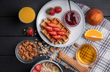 Breakfast. Morning meal set. Delicious oatmeal granola with berries, nuts and honey. A crispy waffle topped with strawberry jam. On a black table background. Top view