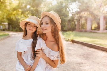 Inspired young woman with beautiful tan posing with pleasure near little daughter in stylish...