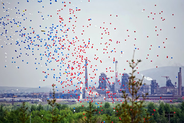 Many multicolored balloons in flight against the background of the pipes of a large metallurgical plant. Contrasts of nature, industrial enterprises and the results of human activity