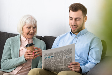 Fototapeta na wymiar smiling senior mother holding coffee and reading newspaper with son at home