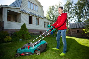 Young man mowing the lawn. Worker doing his job in backyard. Spending summer day in garden. Lawnmower standing on the background of private    wooden house cottage.