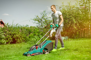 Young man mowing the lawn. Worker doing his job in backyard. Spending summer day in garden. Lawnmower standing on the background of private garden.