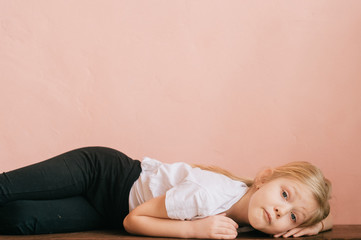 Lonely little girl with pensive tranquil face lying on wooden surface at home and looking at camera with deep emotional eyes.  Lifestyle portrait of poor offensed female child on pink wall background.