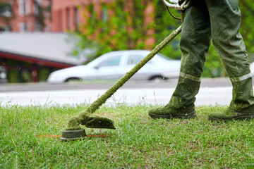 Worker mowing lawn with grass trimmer roadside on sunny day. Professional service gardener using trimmer. Worker cutting grass in green field with lawn mower