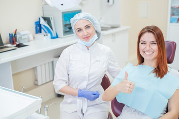 A female dentist and a red-haired patient in an office after dental treatment are looking at the camera with happy faces. Happy woman with her doctor after taking shows sign for shelter.