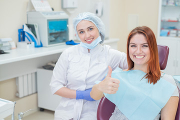 Happy women a dentist and patient after treating teeth at the dental office, smiling and looking towards the camera. Happy patient shows thumb as an endorsement. Dental equipment.