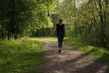 Fototapeta premium A young girl walks in the park. Spring, warm day. A girl with a good figure, blonde, in jeans and a light blouse. The trees have already dissolved the leaves. Falling shadow from the trees.