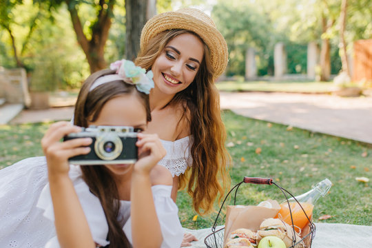 Portrait of laughing long-haired lady in hat with girl holding camera on foreground. Outdoor photo of young woman having fun at picnic and her daughter sitting besie basket with meal.