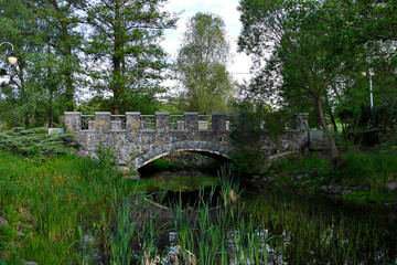 Fototapeta na wymiar Stone bridge over the creek. flowing among large green trees and bushes. Early summer morning.
