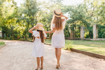 Tanned girl and young woman standing on tip-toe, holding hands and looking at distance. Full-length portrait of slim mother and long-haired daughter in park wears similar straw hats.