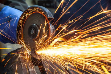 Close-up on the sides fly bright sparks from the angle grinder machine. A young male welder in a white working gloves grinds a metal product with angle grinder in the garage - obrazy, fototapety, plakaty