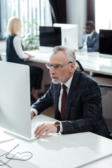 selective focus of handsome mature businessman in glasses looking at computer monitor near multicultural colleagues in office