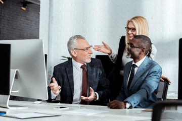 cheerful blonde woman gesturing while looking at businessman near african american partner