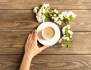 A cup of hot aromatic coffee in female hands and a blooming apple tree branch on a wooden background. Flat layout.