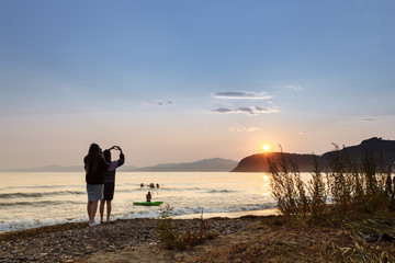 Two girls standing by the seaside. A heart sign. Waves and  beautiful sunset. 
