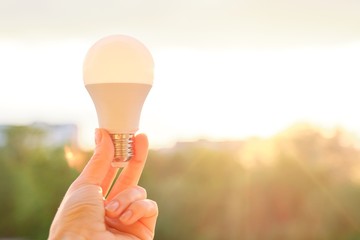 Led bulb, hand holding lamp, evening sunset sky background
