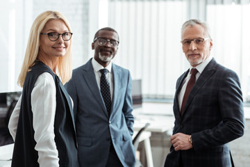 selective focus of cheerful businesswoman in glasses looking at camera near multicultural partners