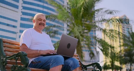 Senior man in a white t-shirt sitting in the Park on a bench among the palm trees and looking at the laptop screen - Powered by Adobe